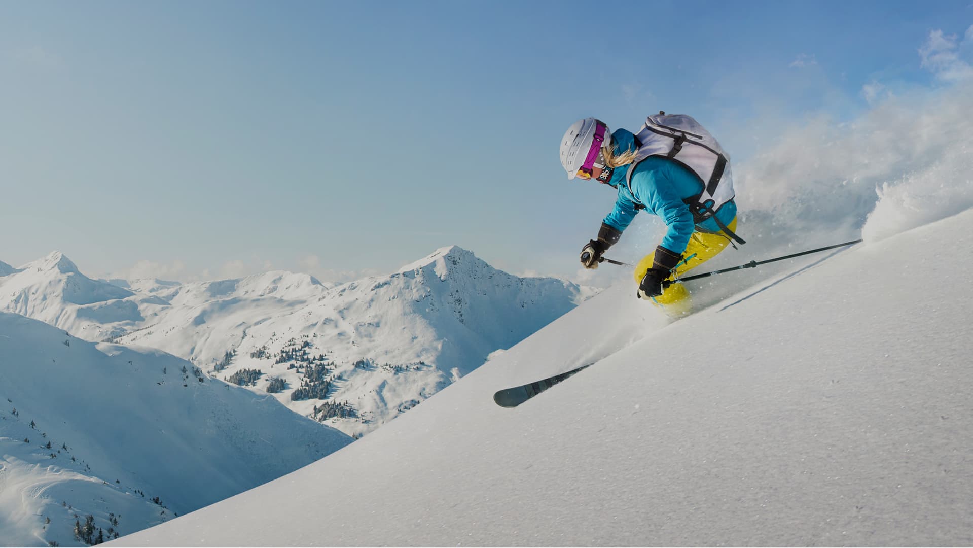 A photo of a skier wearing a black helmet, green jacket and yellow trousers skiing through a tree covered mountain